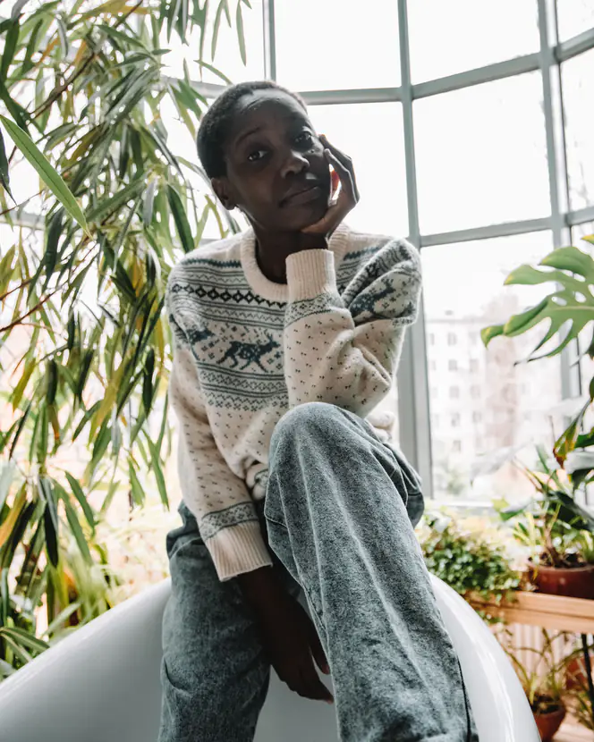 A young woman in a knitted sweater sitting among indoor plants, looking out of a window.