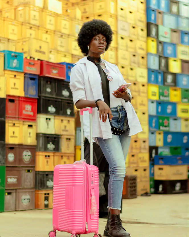 A young woman with a pink suitcase standing against a wall of colourful crates.