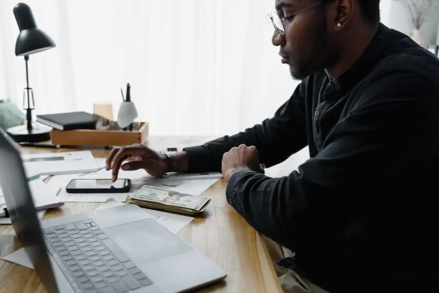 A man at a desk with a laptop, phone and United States dollar notes.